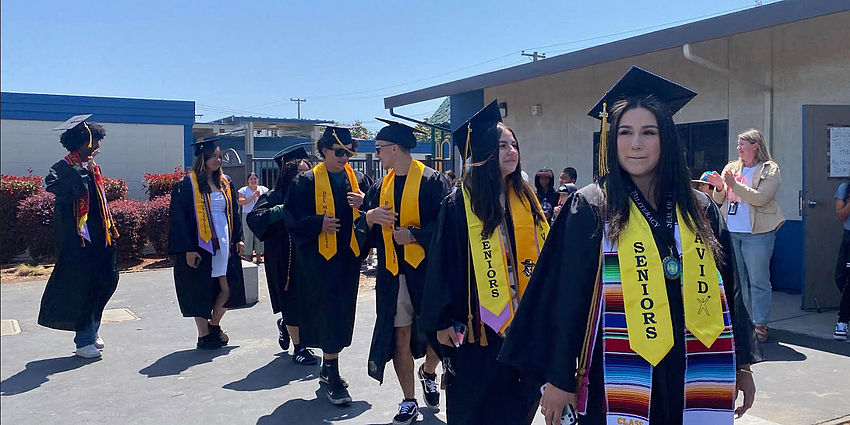 high school grads in caps and gowns walk in line through elementary campus