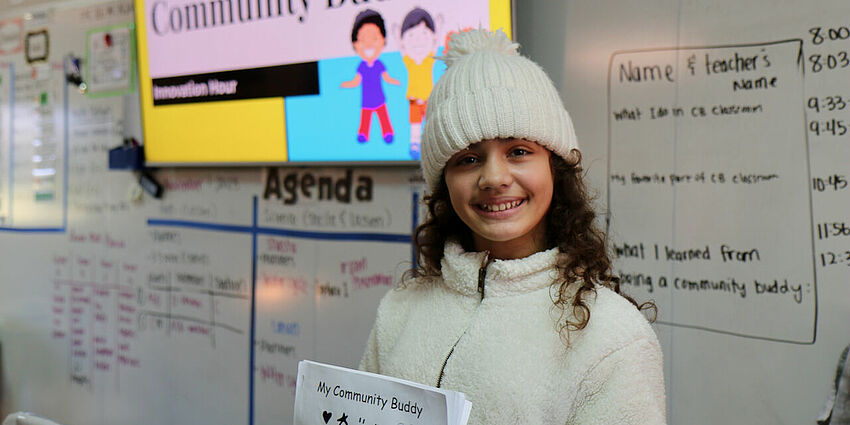 one girl smiling and wearing a knitted cap. Behind her is a large screen that says Community Buddies