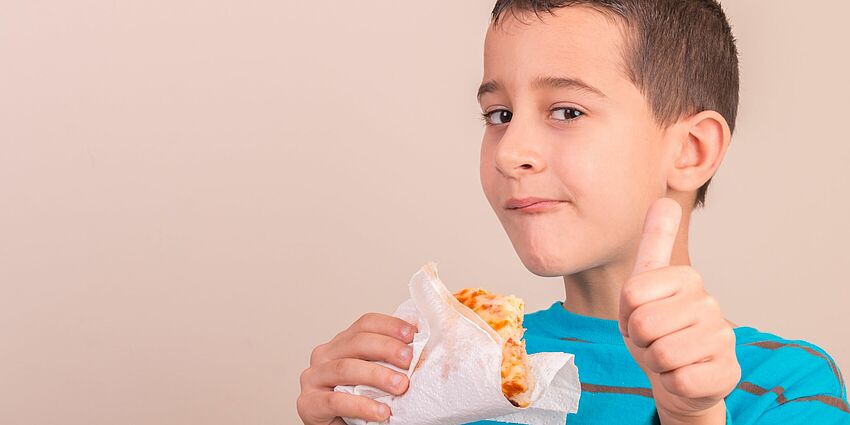 boy holding a slice of pizza in one hand and giving a Thumbs Up with the other.