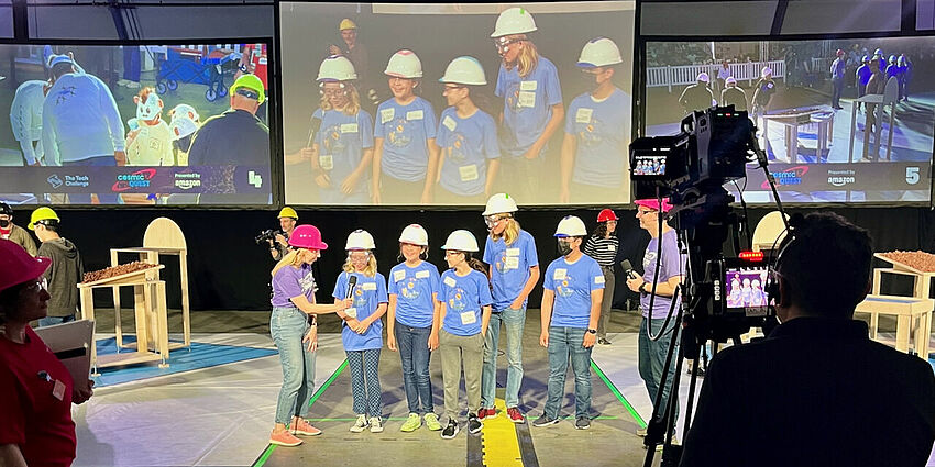 5 students in white hard hats standing in front of cameras as a woman in a pink hard hat holds a microphone and a man in a pink hard hat listens to what is being said.