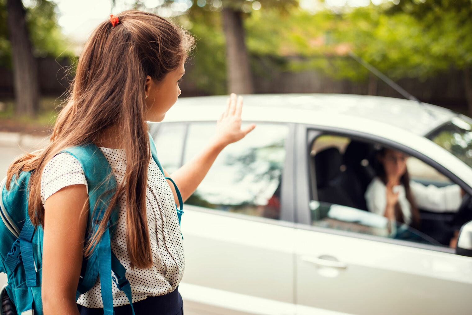 girl with backpack is dropped off at school and waves goodbye to parent in car